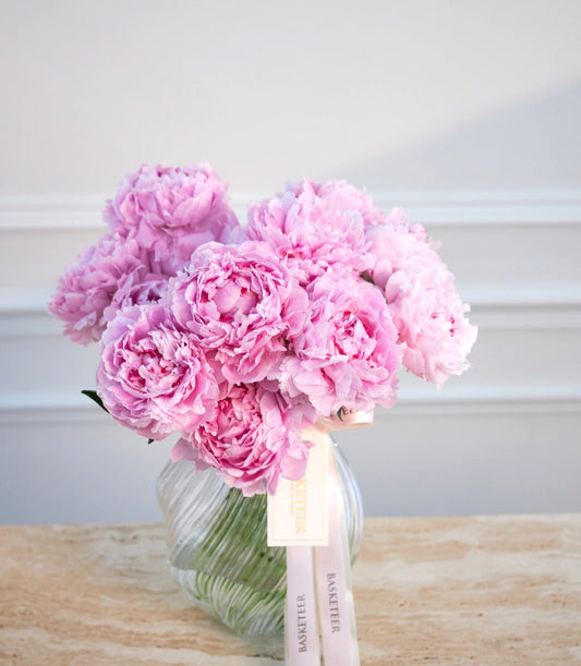 Pink peonies in clear glass vase with green stems and white ribbon bow on wooden surface against white wall.