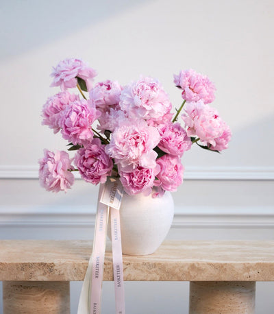 Pink peonies in a white ceramic vase with a pale pink ribbon bow on a wooden shelf against a white wall.