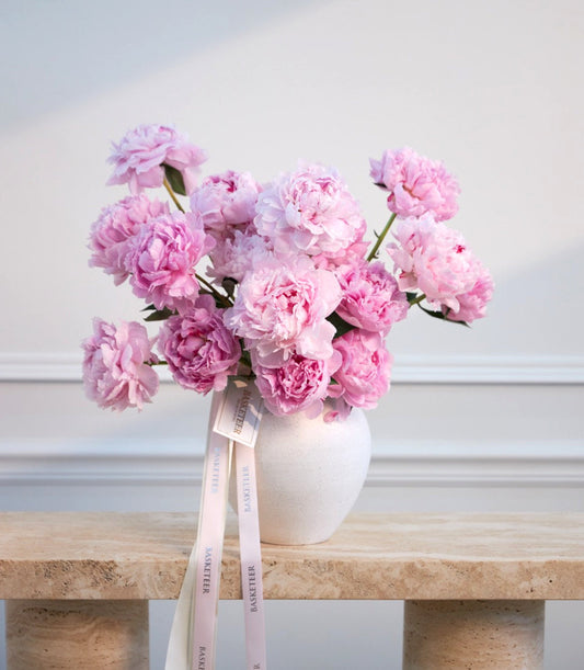 Pink peonies in a white ceramic vase with a pale pink ribbon bow on a wooden shelf against a white wall.
