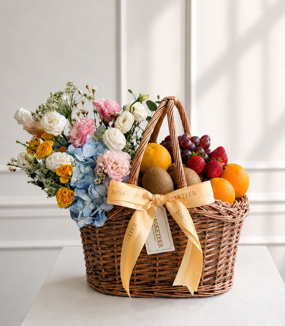 Wicker basket with fresh flowers, fruits, and berries tied with gold ribbon. Contains white, pink, yellow roses, blue hydrangeas, oranges, raspberries, and grapes.