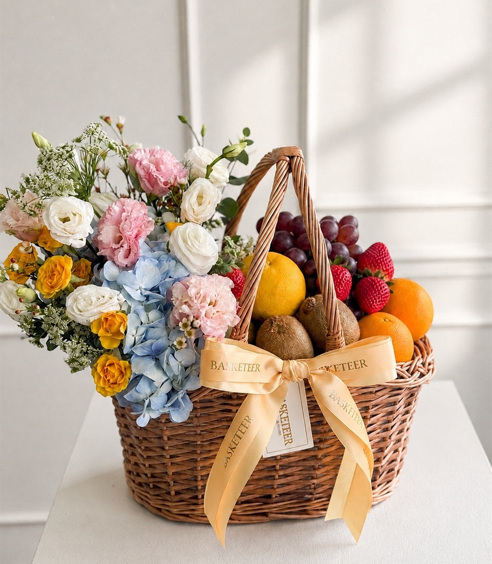 Wicker gift basket with mixed flowers (white roses, pink carnations, yellow roses, blue hydrangeas) and fresh fruits (grapes, raspberries, oranges, kiwis, lemon) tied with peach ribbon.