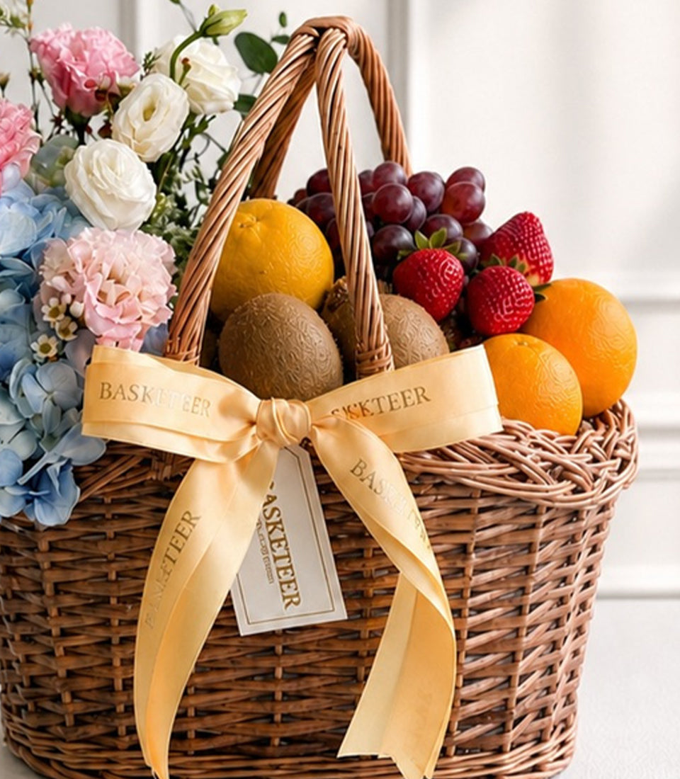 Woven wicker basket filled with fresh fruits, flowers, and a cream ribbon bow, featuring oranges, grapes, strawberries, and kiwis beside pink and white blooms.