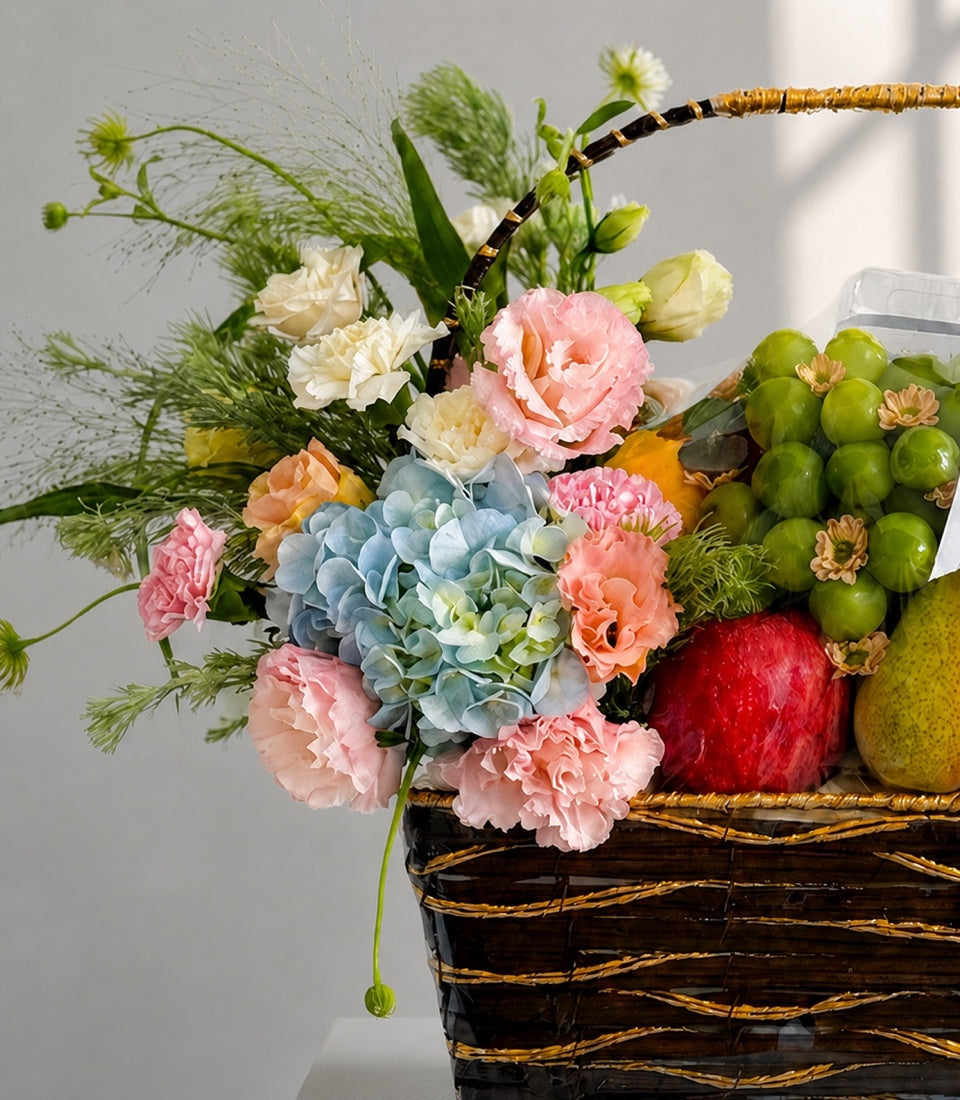 Elegant wicker basket arrangement with pink and cream carnations, blue hydrangea, fresh fruits including red apples and green grapes, adorned with golden and black ribbon.