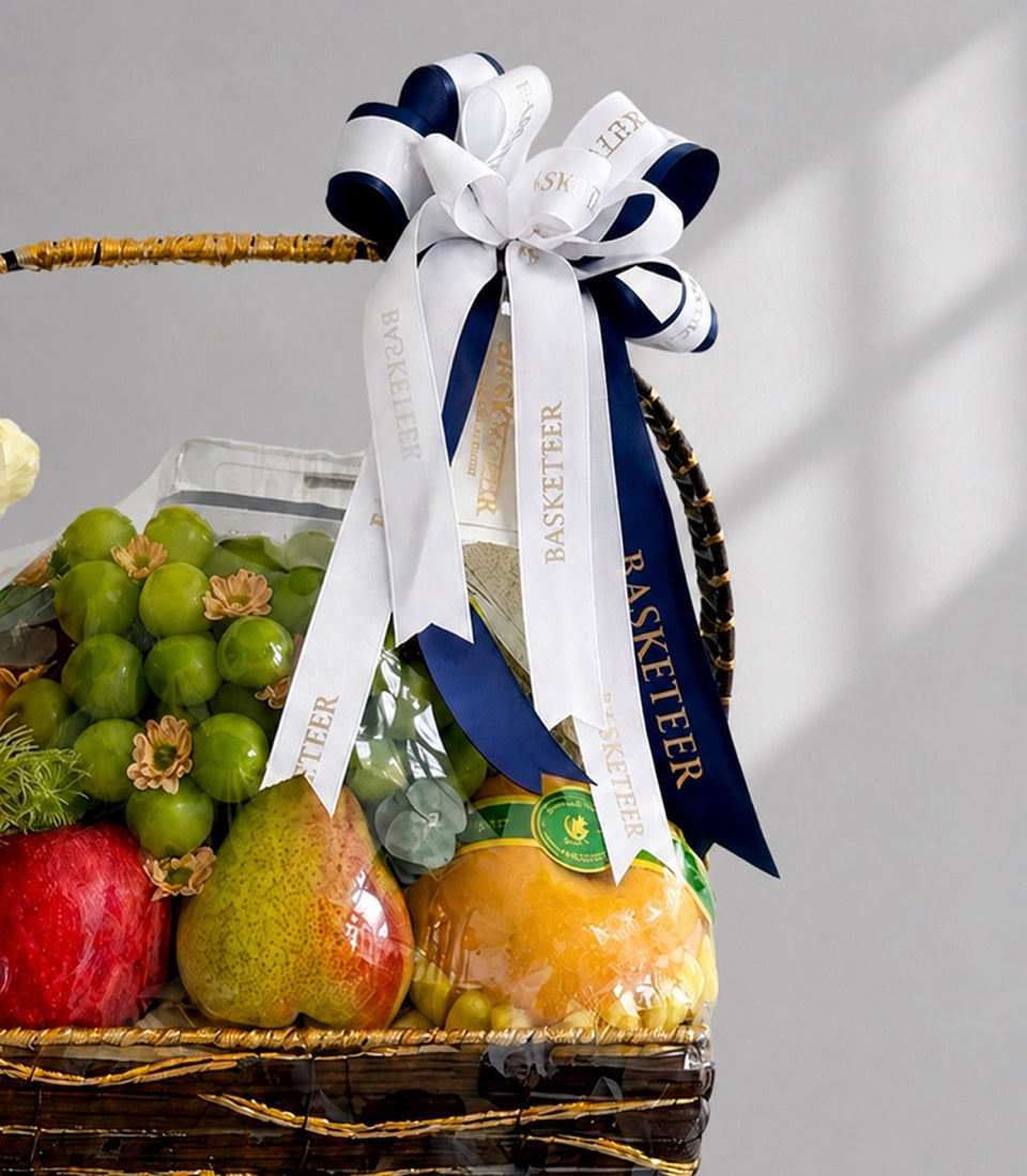 Luxury fruit basket with gold-wrapped handle, navy and white ribbons, containing green grapes, red apple, pears, oranges, and flowers against white background.