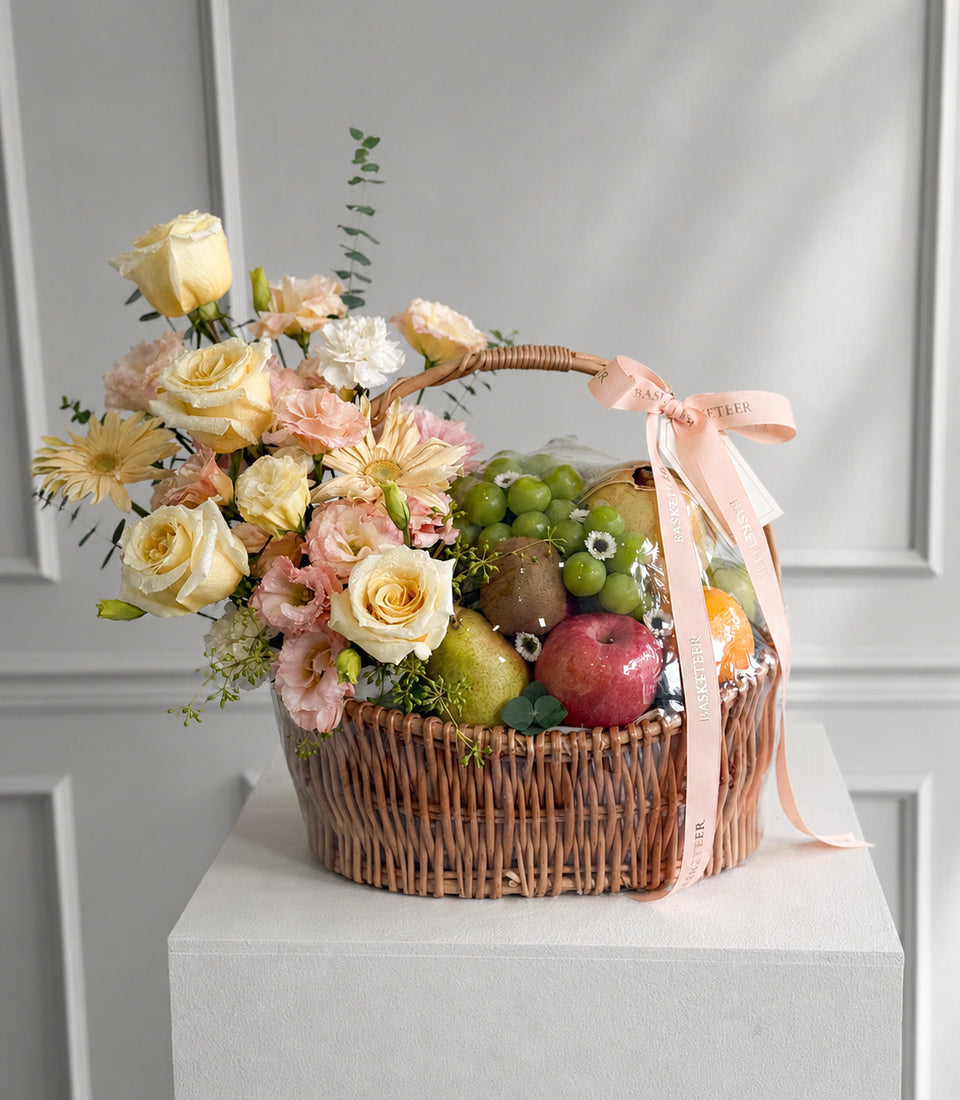 Elegant wicker basket with cream and pink roses, white daisies, fresh fruits, and eucalyptus greenery, tied with a blush ribbon bow.