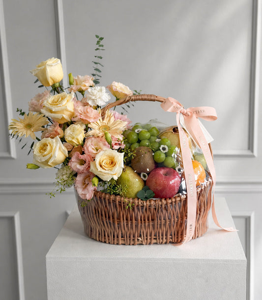Elegant wicker basket with cream and pink roses, white daisies, fresh fruits, and eucalyptus greenery, tied with a blush ribbon bow.