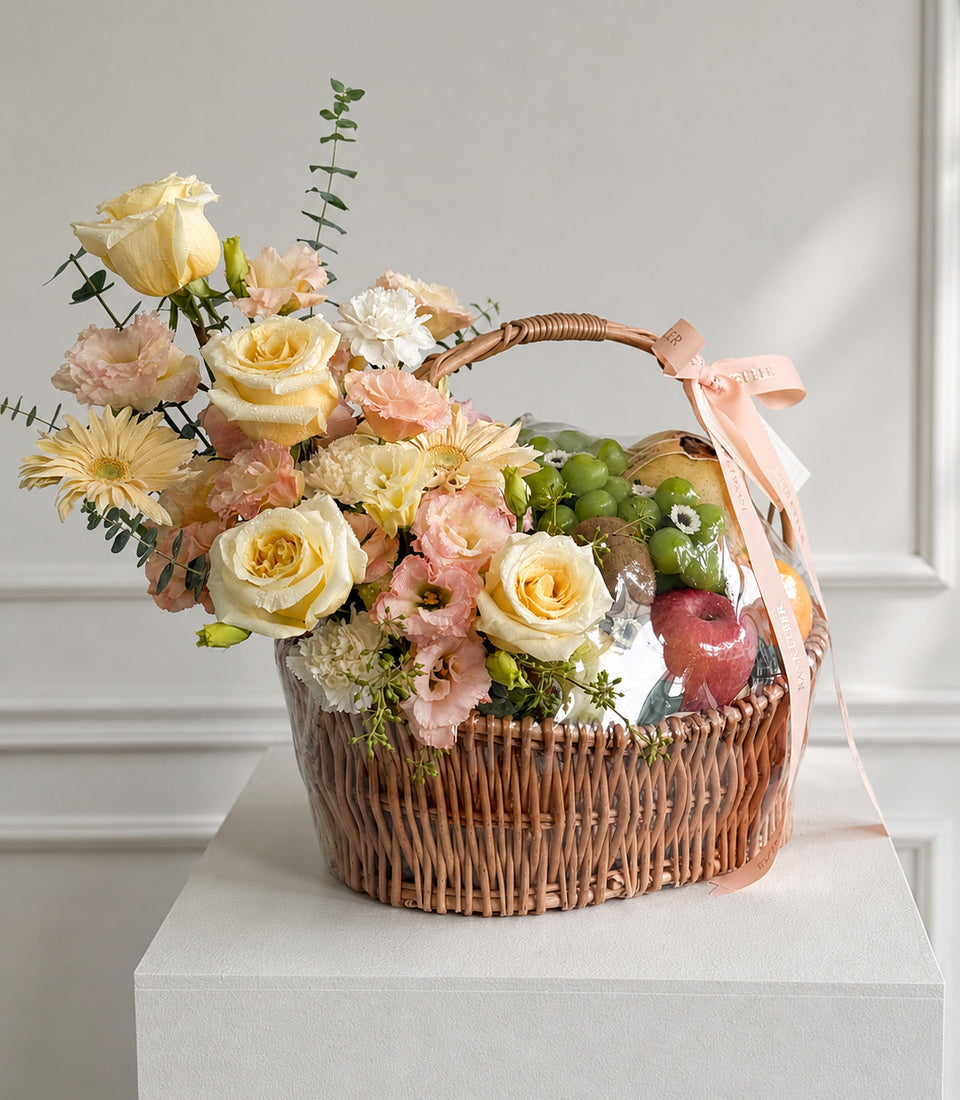 Wicker basket filled with yellow roses, pink carnations, cream flowers, green berries, and red apple, tied with blush ribbon.