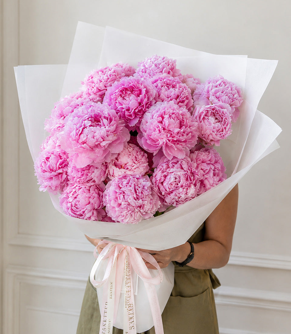 Luxurious bouquet of pink peonies wrapped in white paper with pink ribbon, held by person in olive dress against neutral background.