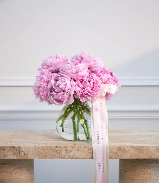 Pink peonies in a clear glass vase with green stems, decorated with a pale pink ribbon bow, displayed on a wooden shelf against a white wall.