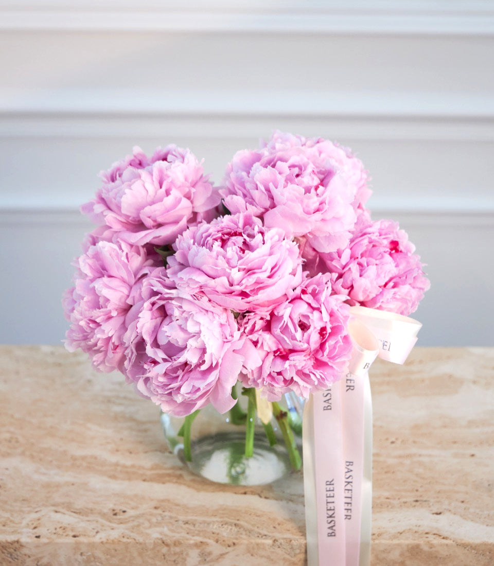 Pink peonies in clear glass vase with green stems, tied with pale pink ribbon labeled "Basketeer," on wooden surface.