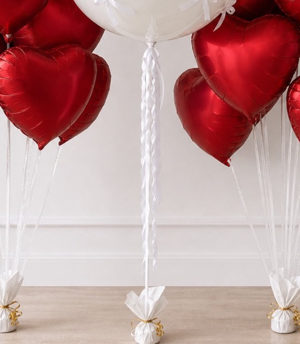 Red heart-shaped foil balloons with white ribbons and gold-tied balloon weights on a neutral background.