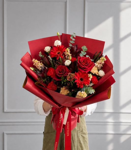 A vibrant red rose bouquet with gerbera daisies, white spray roses, and lush