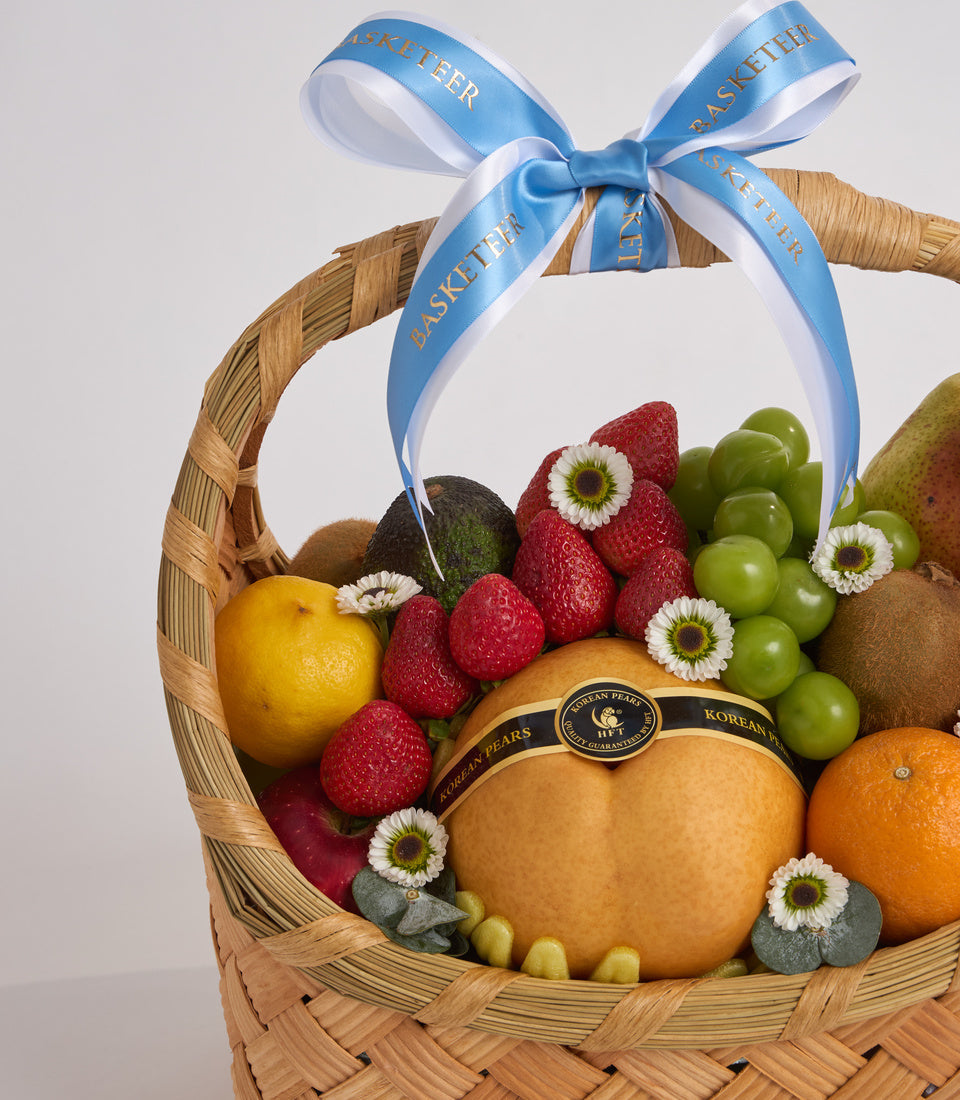 Woven wicker basket filled with fresh fruits including strawberries, grapes, oranges, and pears, decorated with white flowers and tied with a blue ribbon bow.