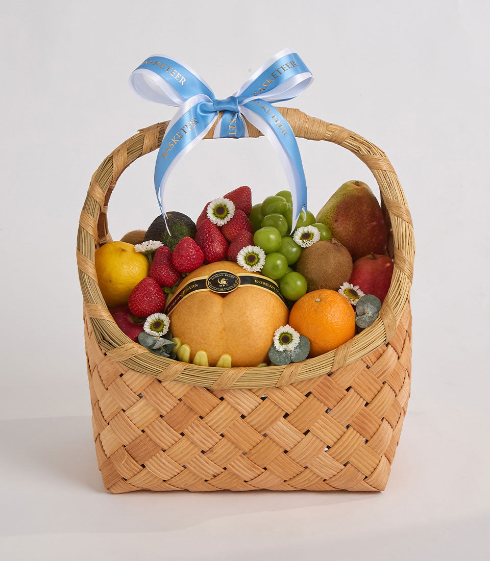 Woven wicker fruit basket with blue ribbon bow, filled with raspberries, grapes, pears, oranges, lemons, and white daisy flowers.