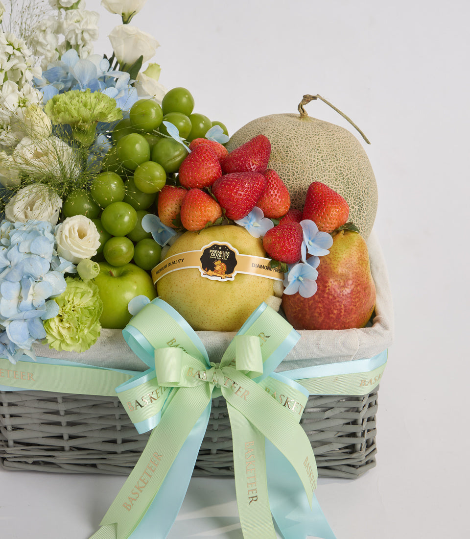 Luxury fruit and flower gift basket with green grapes, strawberries, pear, melon, white roses, blue hydrangeas, and a green ribbon bow.