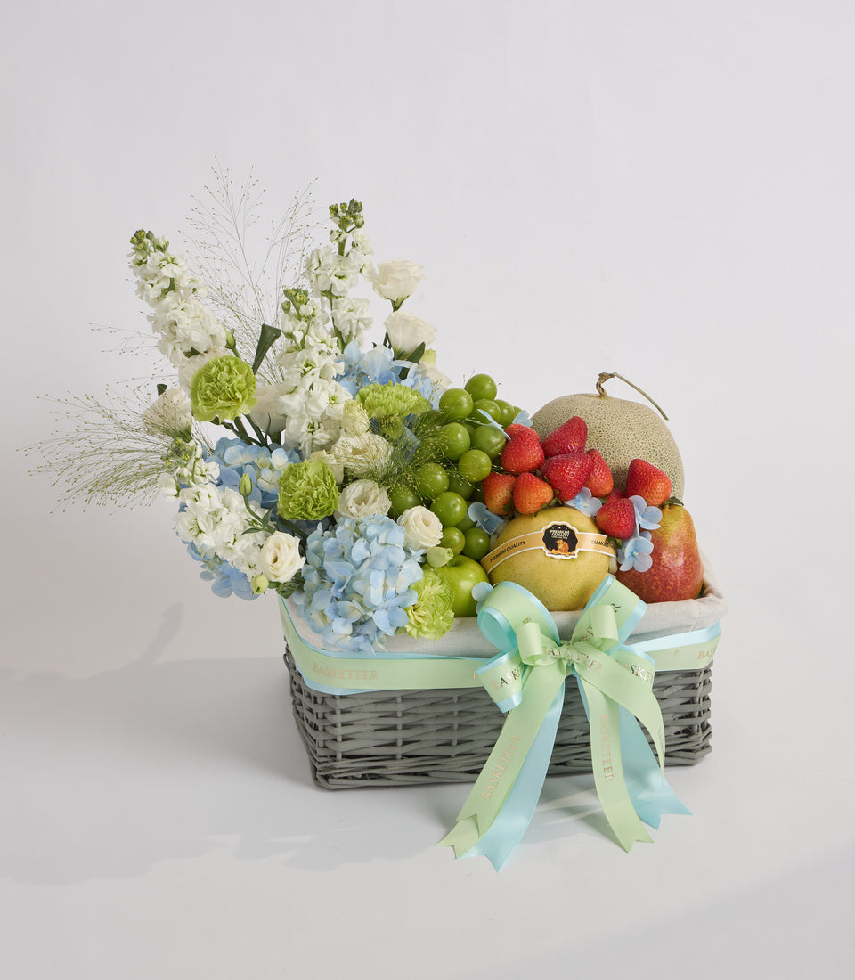 # Alt Text:

Gray wicker basket with green and blue ribbon bow containing white and blue flowers, fresh raspberries, green grapes, and melons on white background.