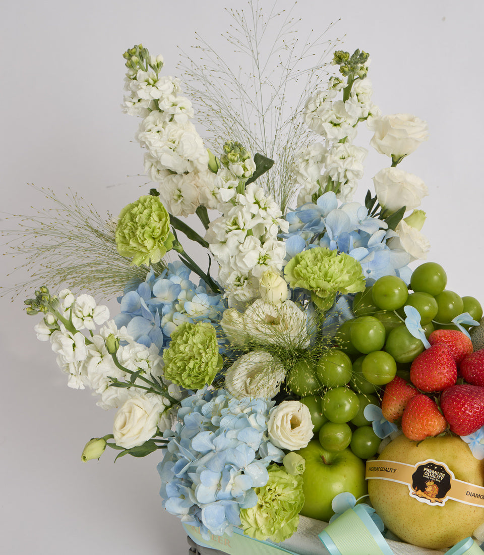 Luxurious floral and fruit arrangement featuring white stocks, blue hydrangeas, green chrysanthemums, ornamental grasses, fresh strawberries, green grapes, and pears on white background.