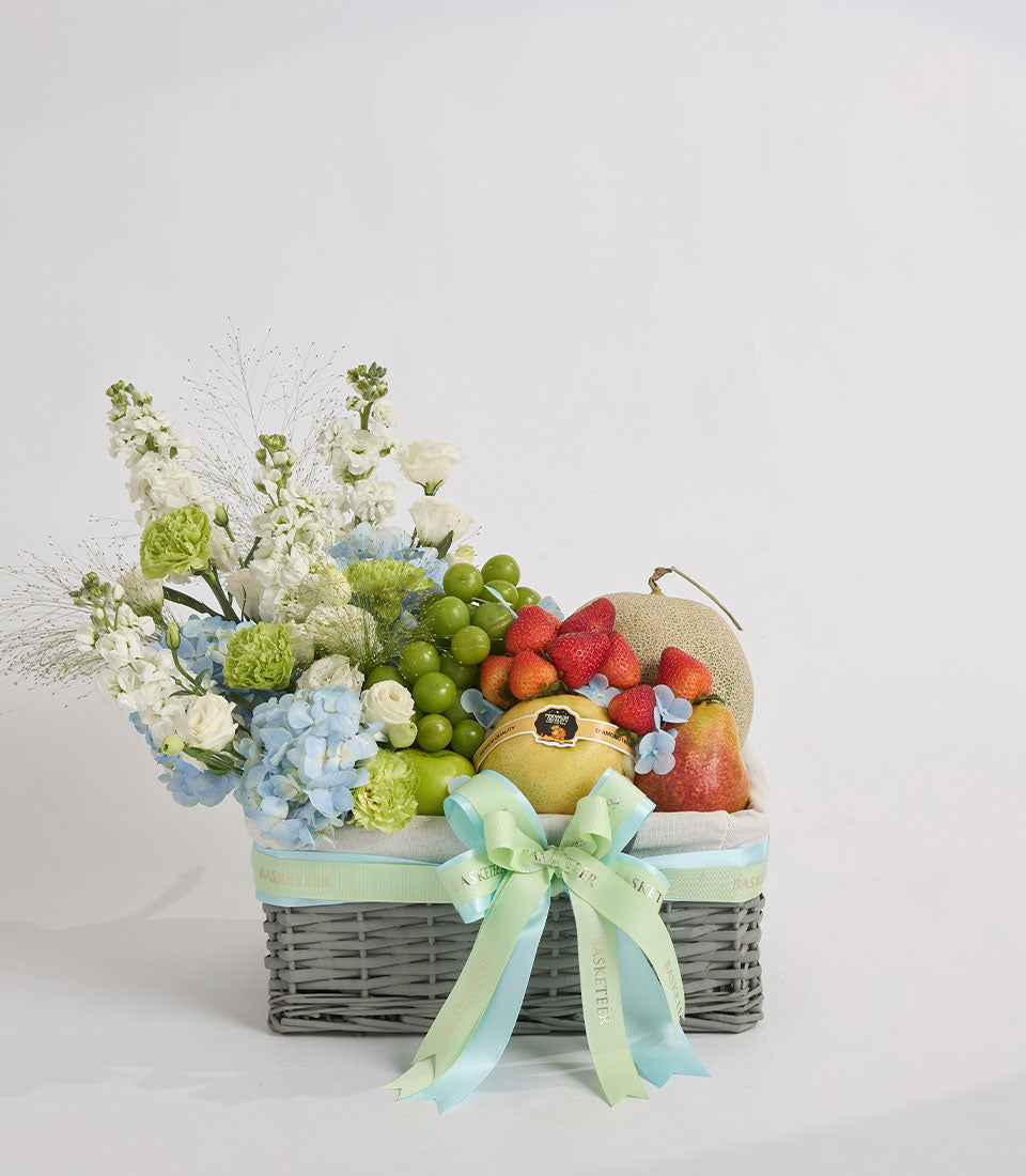Gray wicker basket with fresh flowers, fruits, and a decorative ribbon bow. Contains white roses, blue hydrangeas, green carnations, pears, strawberries, and grapes.