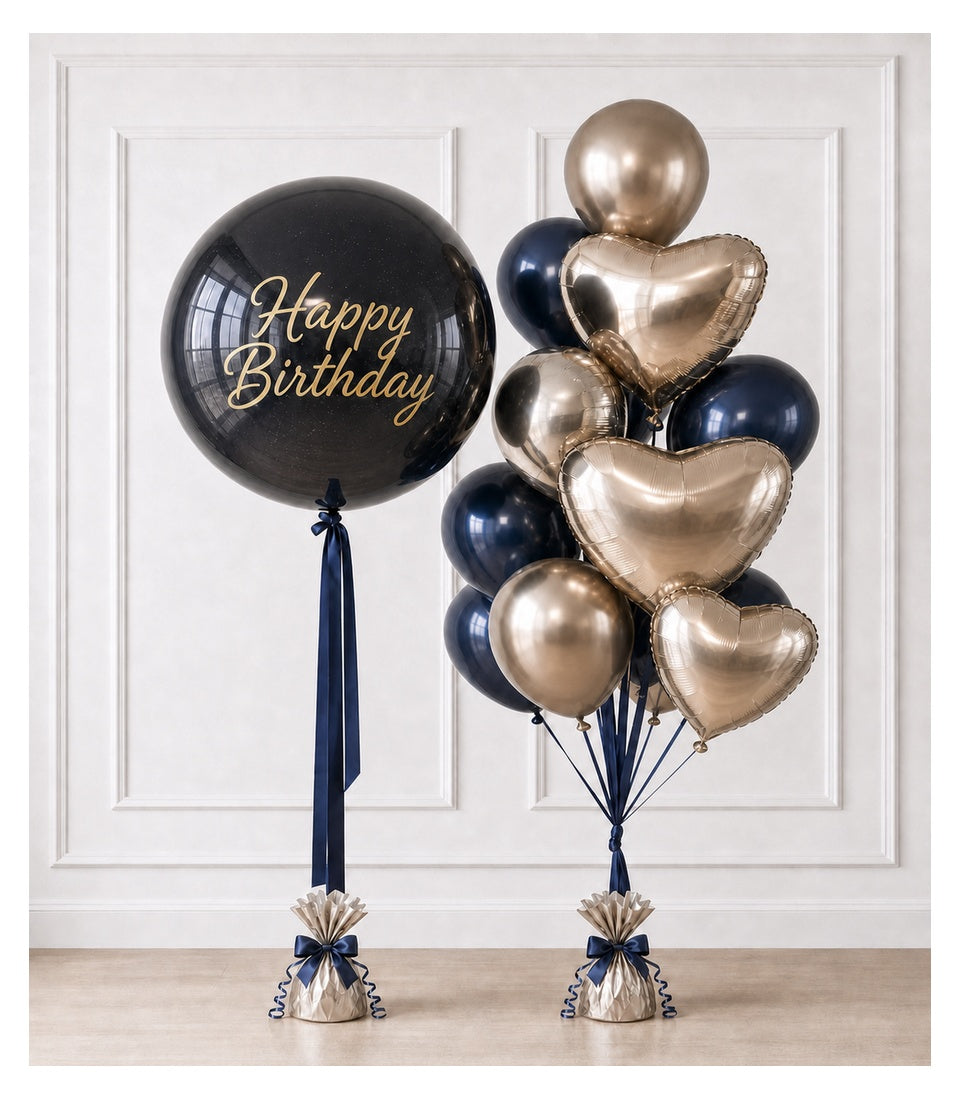 Black and gold "Happy Birthday" balloon bouquet with heart-shaped and round foil balloons, navy ribbons, and weighted bases against white paneled wall.