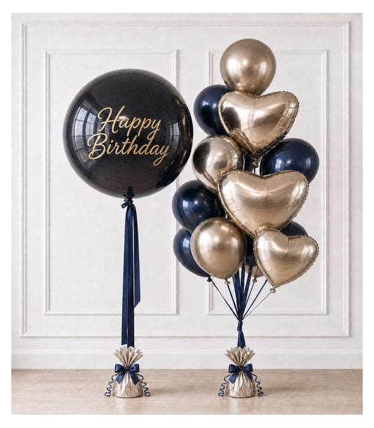 Black and gold "Happy Birthday" balloon bouquet with heart-shaped and round foil balloons, navy ribbons, and weighted bases against white paneled wall.