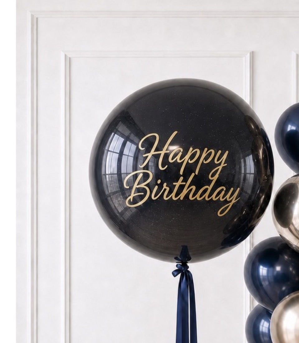 Black orb balloon with gold "Happy Birthday" text and navy ribbon bow, displayed against white paneled wall with matching metallic balloons.