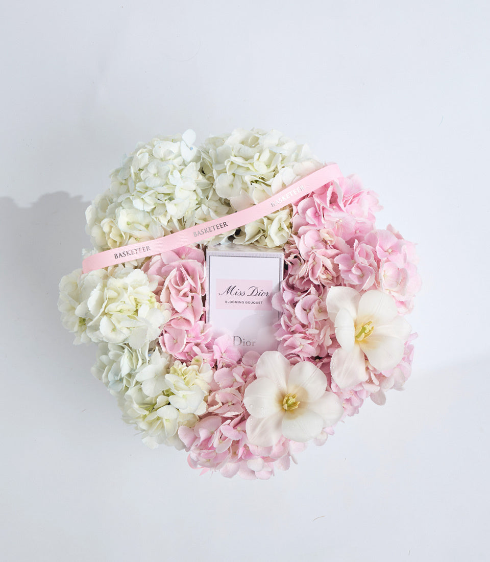 Miss Dior Blooming Bouquet perfume surrounded by white hydrangeas, pink peonies, white orchids, and pink ribbon on white background.