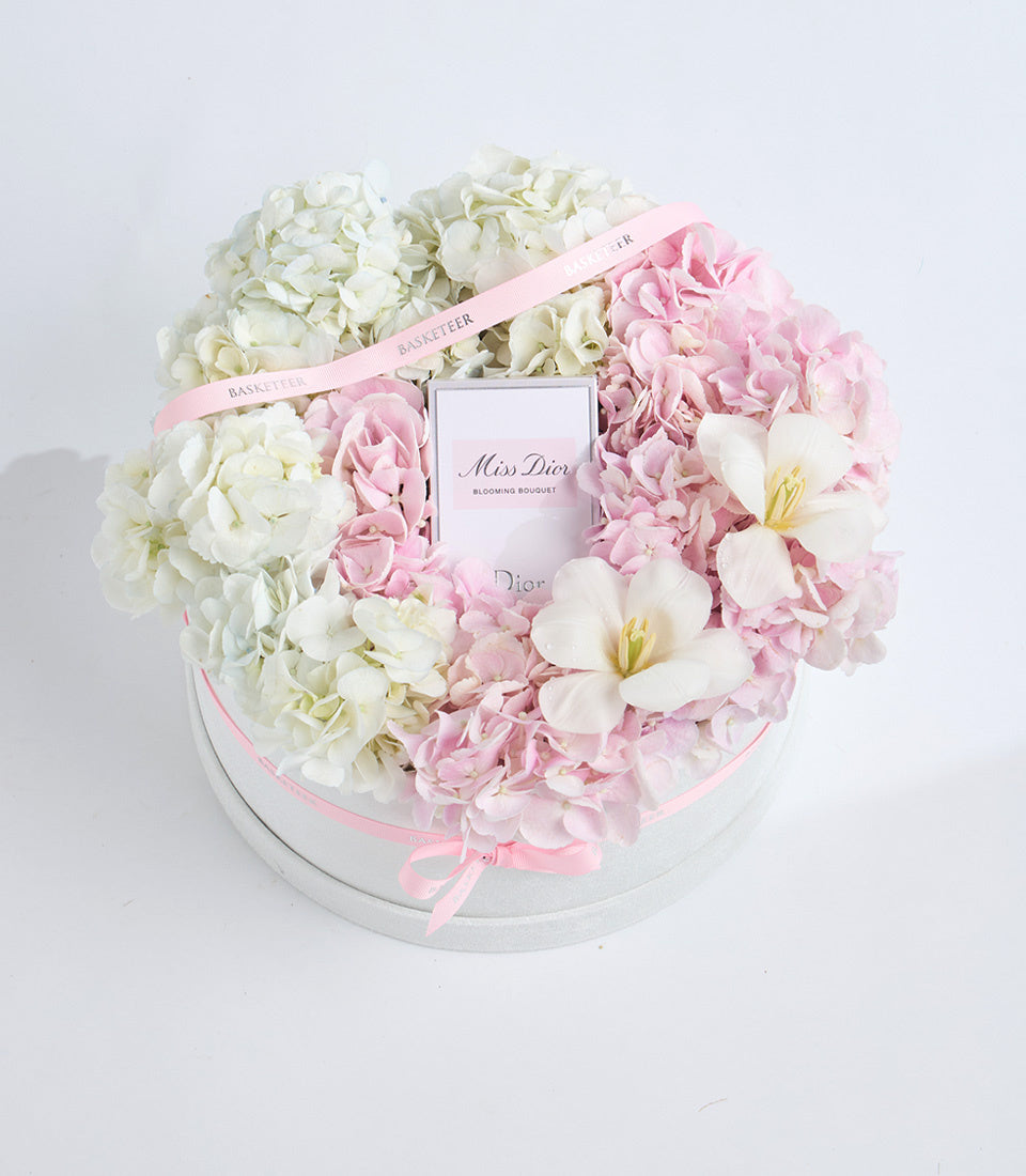 Luxurious floral arrangement featuring white hydrangeas and pink peonies with white flowers in a white box, centered with Miss Dior perfume bottle and pink ribbons.