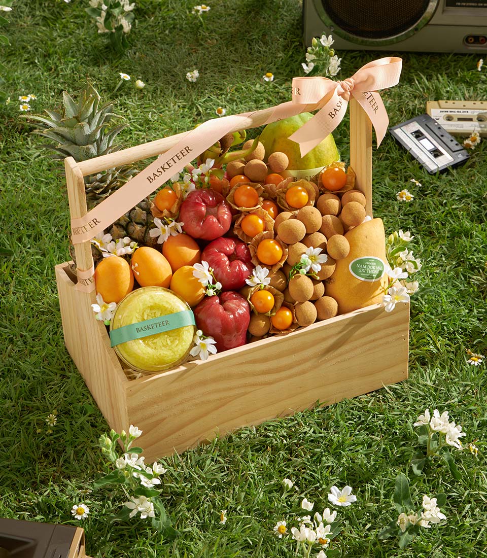 Wooden basket filled with fresh Thai tropical fruits including rose apples, mangoes, longans, and pineapple with pink