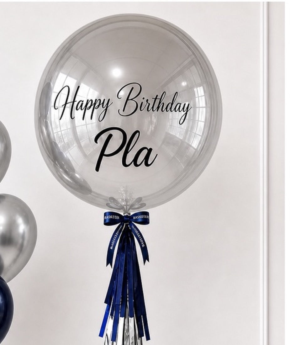 Clear bubble balloon with "Happy Birthday Pla" text, navy blue and silver ribbon tail, elegant gift decoration.