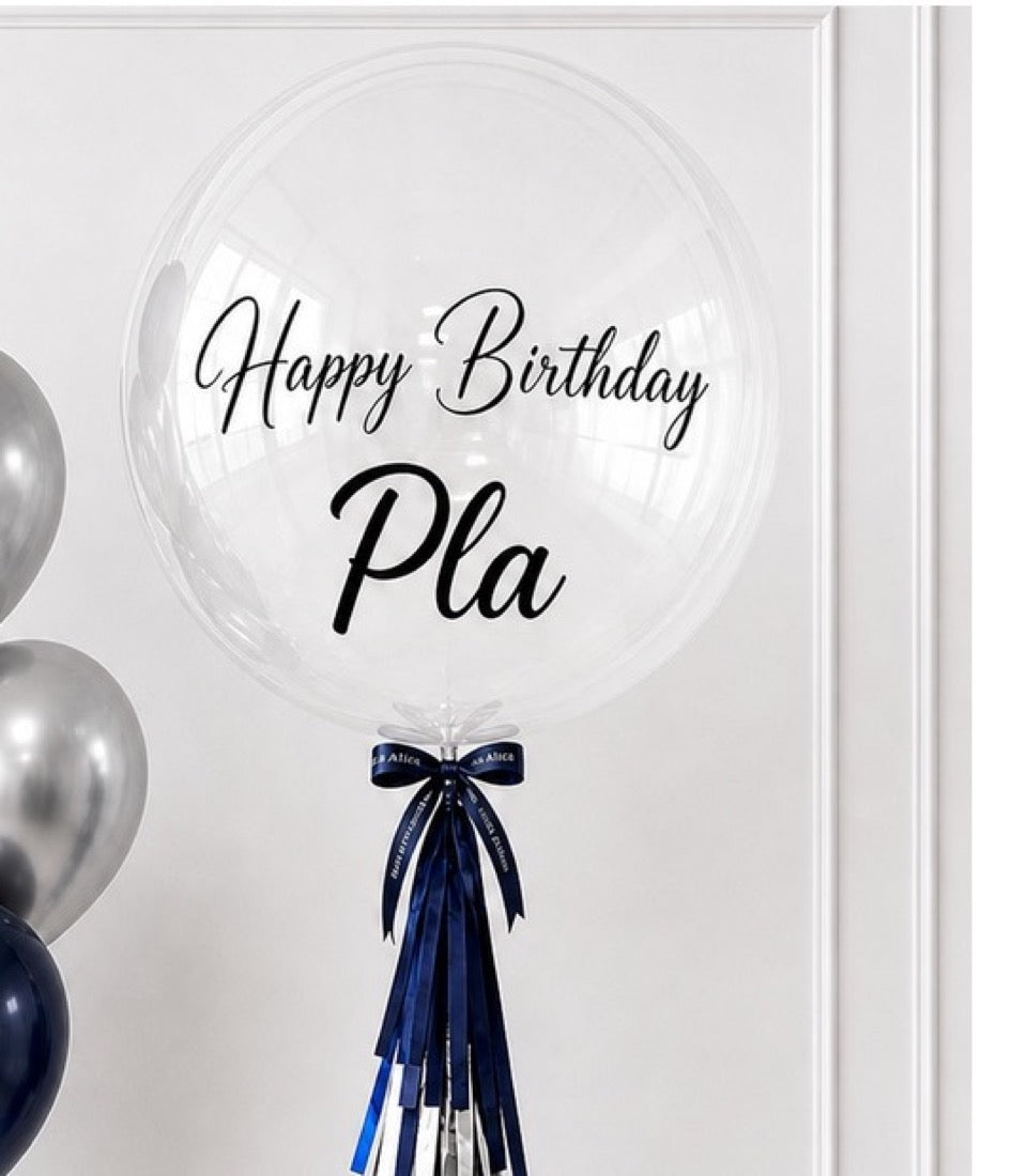 Clear bubble balloon with "Happy Birthday Pla" text, navy blue ribbon bow, silver and navy balloons beside it, elegant minimalist style.