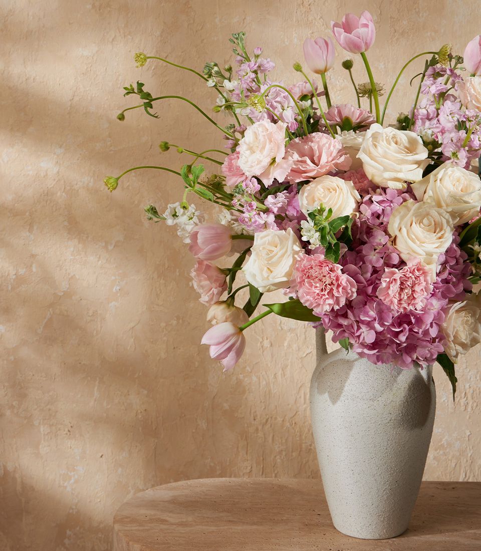 Lush pink and white floral arrangement with tulips, roses, and hydrangeas in textured grey ceramic vase
