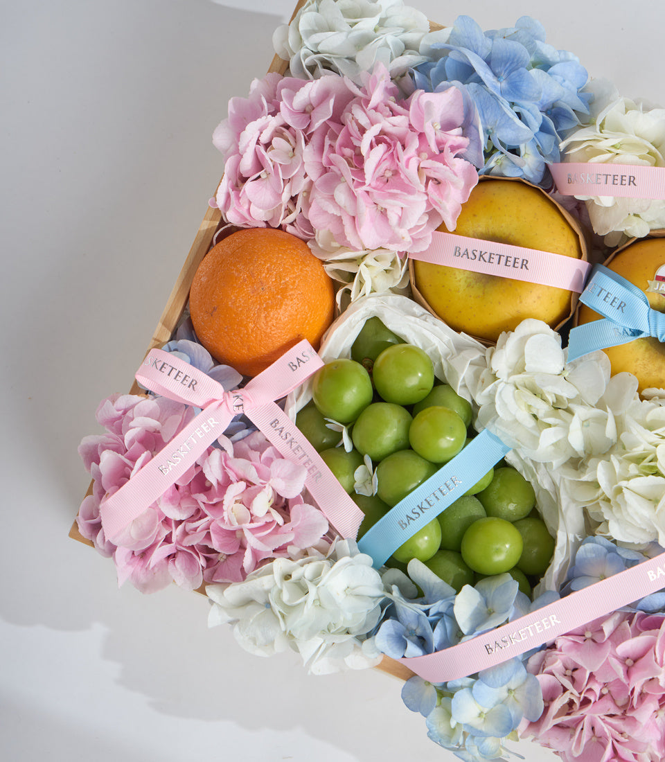 Luxurious fruit and flower gift basket featuring pink hydrangeas, white flowers, blue blooms, fresh oranges, yellow pears, and green apples with pink and blue ribbons.