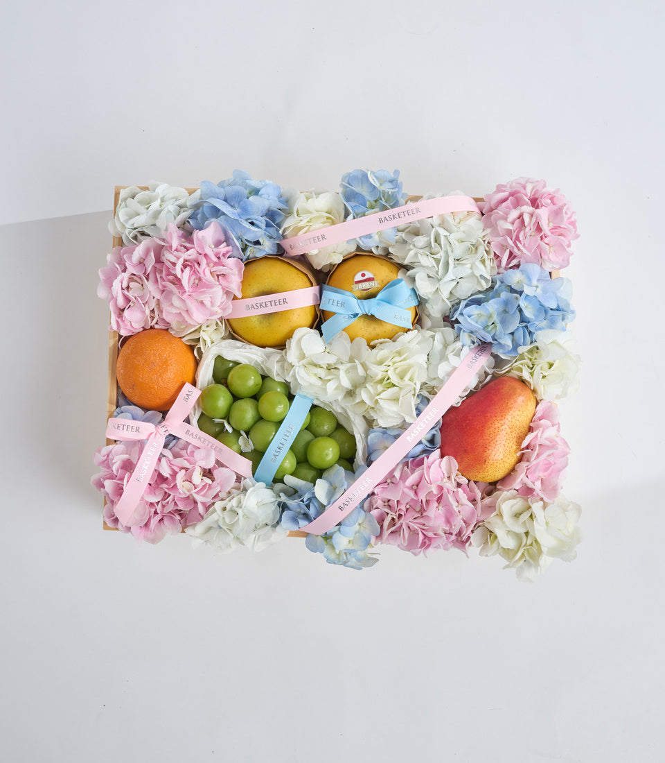 Luxury fruit and flower gift basket with pink carnations, blue hydrangeas, white flowers, fresh oranges, lemons, grapes, and decorative ribbons on natural wood base.