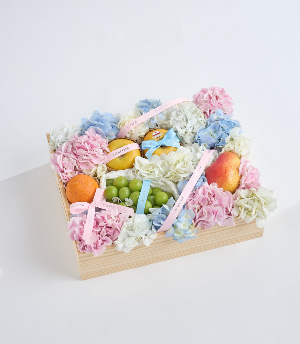 Wooden basket arrangement with pink, blue, and white hydrangeas, fresh fruits (oranges, mangoes, grapes), and decorative ribbons on white background.