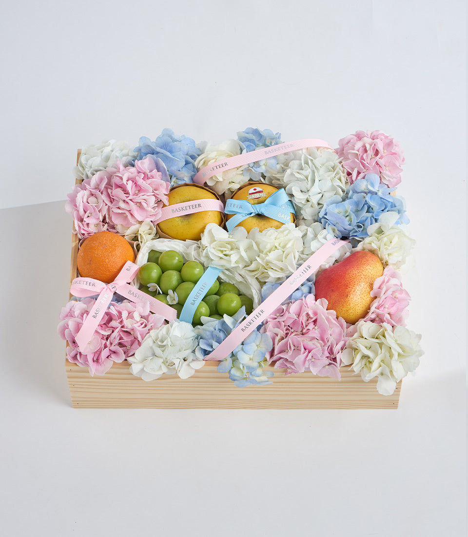 Wooden gift basket with pink and blue hydrangeas, white flowers, fresh fruits including oranges and limes, tied with pastel ribbons.