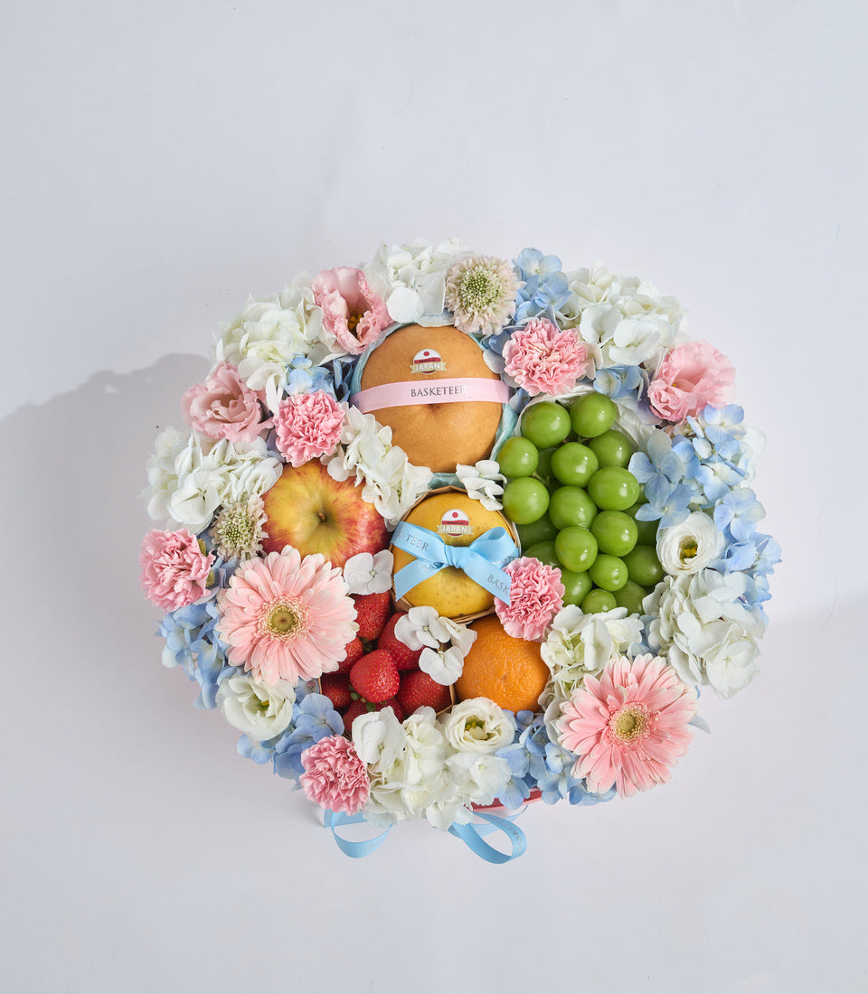 Floral gift basket with pink carnations, white roses, blue hydrangeas, fresh fruits including apples, oranges, green grapes, and raspberries, tied with blue and pink ribbons.