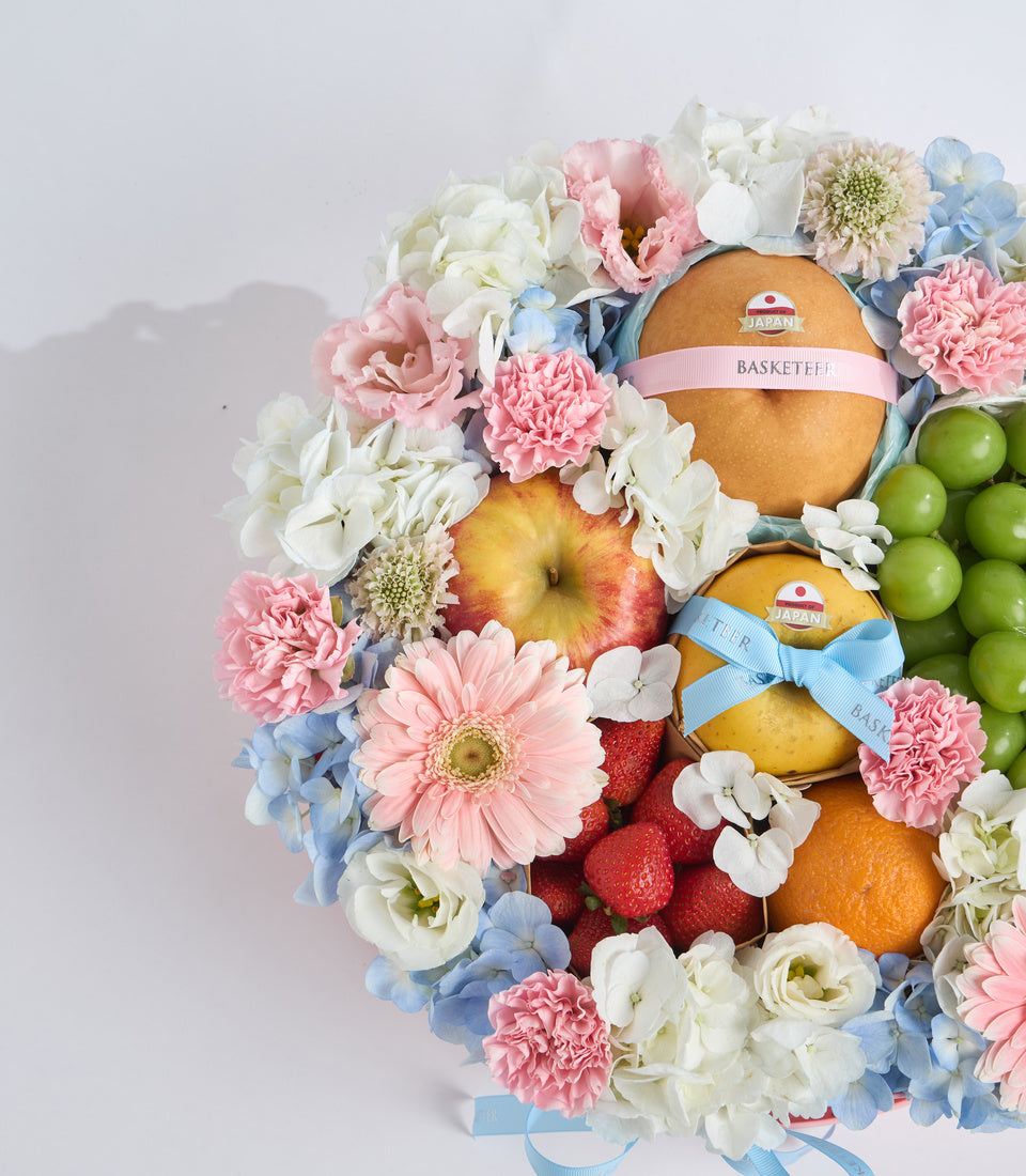 Luxury fruit and flower gift basket with pink carnations, white roses, blue hydrangeas, Japanese pears, apples, strawberries, and green grapes with ribbon bows.