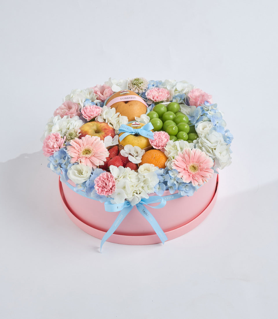 Luxe floral gift box with pink container, featuring pink carnations, white roses, blue hydrangeas, fresh fruits, and pastries with blue ribbon bow.
