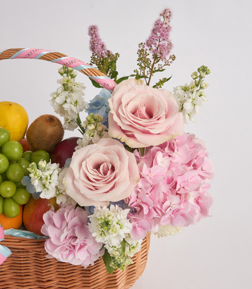 Wicker basket arrangement with pink roses, carnations, white stock flowers, fresh fruits, and decorative ribbon against white background.