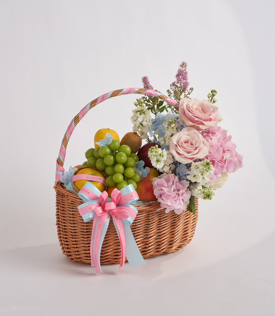 Wicker basket with pink roses, carnations, green grapes, fruits, white flowers, blue hydrangea, and pink ribbon bow on neutral background.