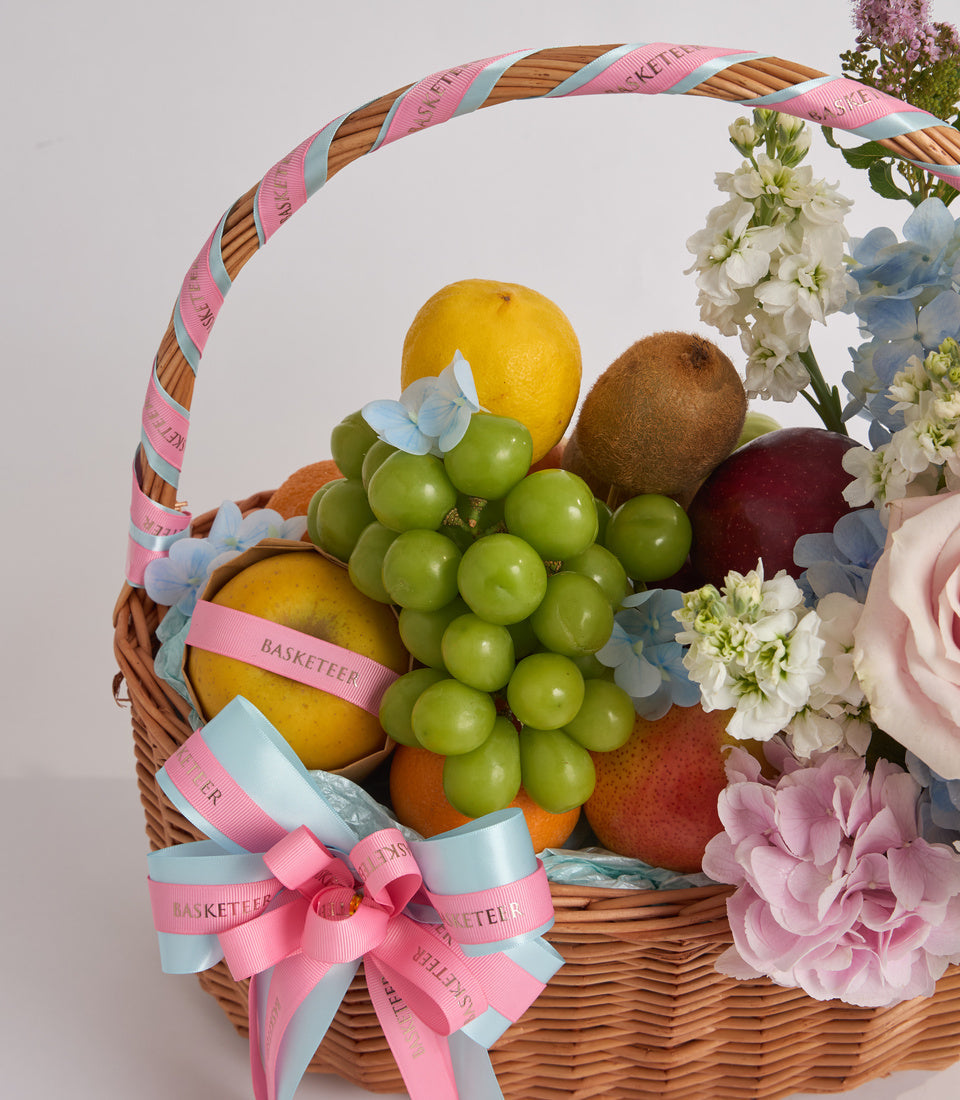 Wicker gift basket with fresh fruits, green grapes, flowers, and pink ribbon bow against white background.