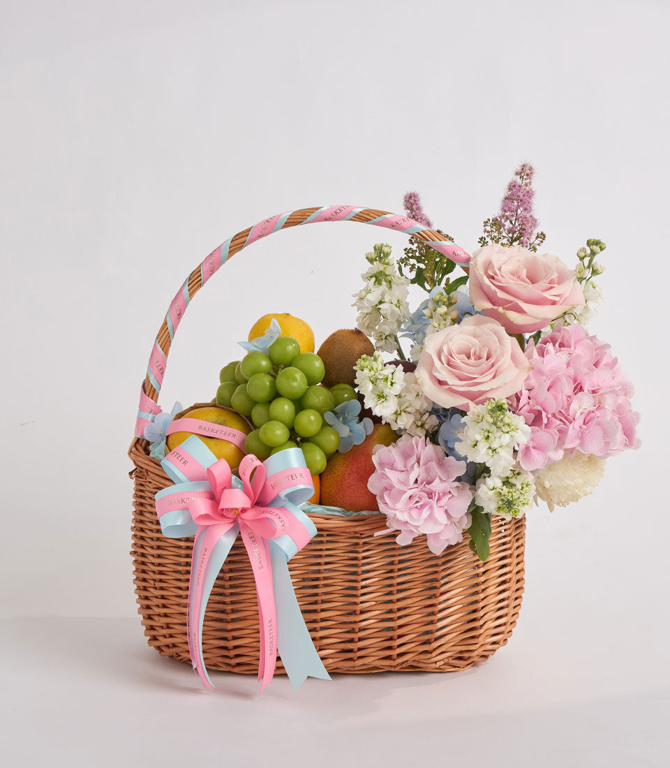 Woven wicker basket filled with pink roses, carnations, white flowers, green grapes, and fresh fruit, tied with a pink and blue ribbon bow.