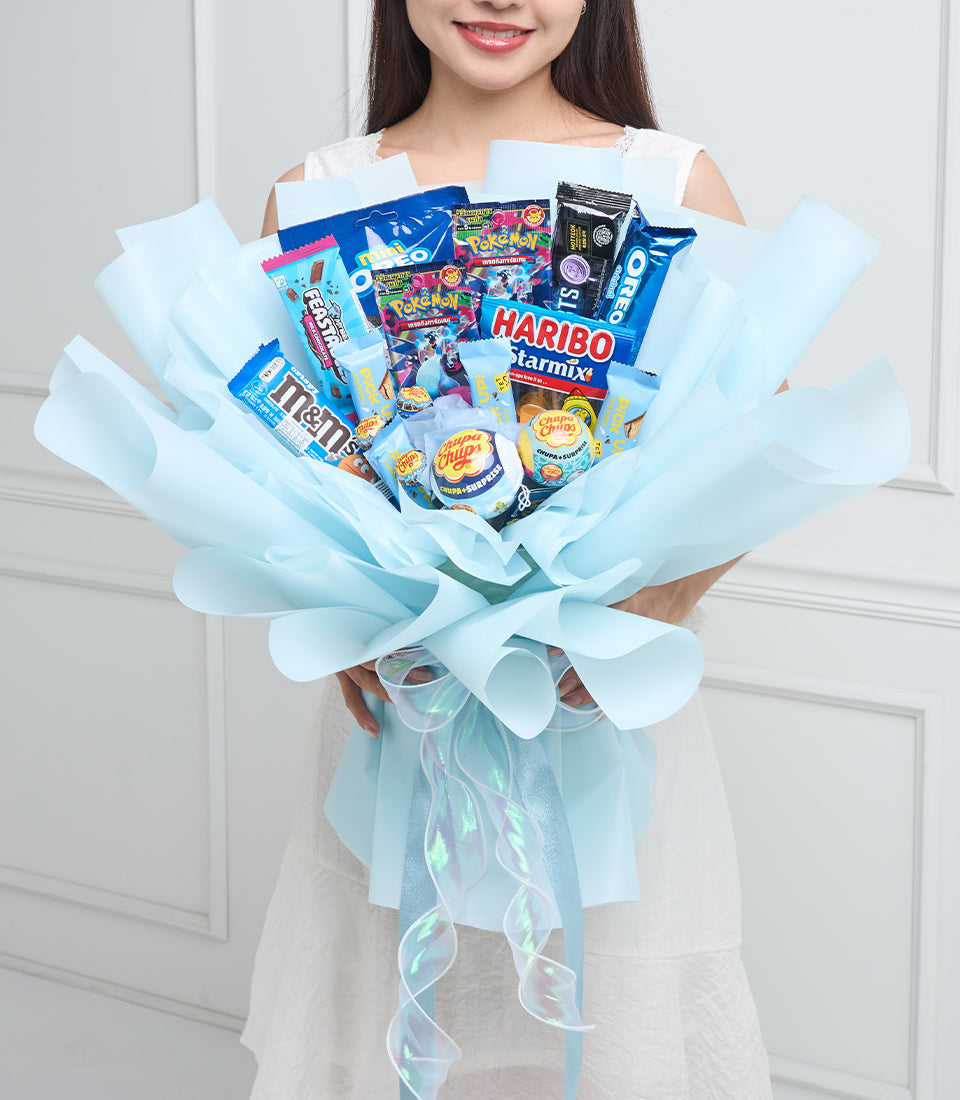 Young woman holding a light blue tissue-wrapped candy bouquet with Pokémon cards, Haribo gummies, M&Ms, Chupa Chups lollipops, and other sweets.