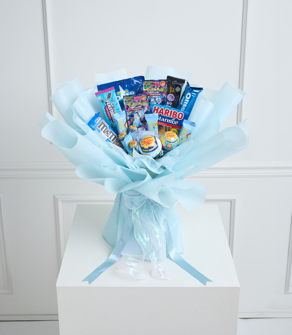 Light blue tissue-wrapped candy bouquet with assorted sweets and snacks including Haribo, M&Ms, and Chupa Chups on white pedestal.