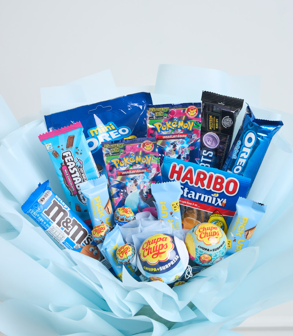 Blue and white gift basket filled with assorted candies, gummies, and snacks including Pokémon cards, Oreo, Haribo, Chupa Chups, and M&M's wrapped in light blue tissue paper.