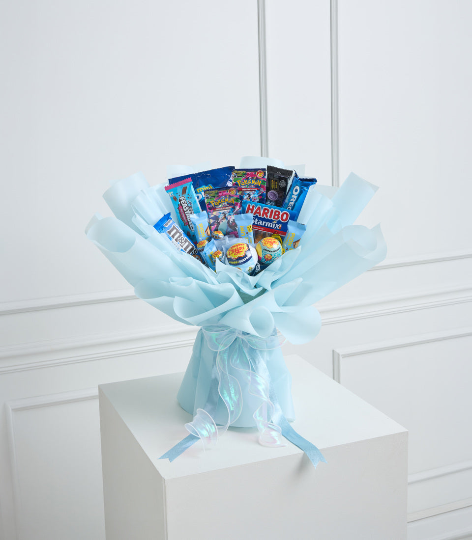 Light blue tissue paper wrapped sweet bouquet with assorted candies including Haribo and Pokemon cards, tied with ribbon on white cube pedestal.