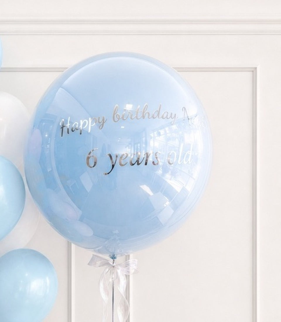 Light blue transparent bubble balloon with "Happy birthday 6 years old" text and white ribbon bow against white wall.
