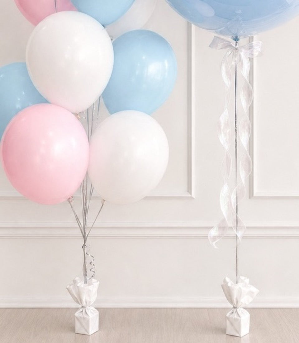 Pastel balloon decorations with pink, blue, and white latex balloons, white ribbon tassels, and weighted gift box bases against a neutral wall.