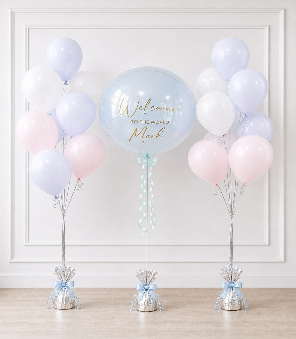 Welcome baby balloon display with large clear bubble centerpiece, pastel pink and lavender balloons, and silver weighted bases with blue bows.
