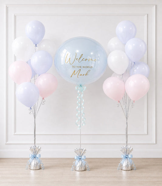 Welcome baby balloon display with large clear bubble centerpiece, pastel pink and lavender balloons, and silver weighted bases with blue bows.